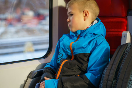 Thoughtful young boy in warm jacket sitting by train window, looking outside during winter travel....