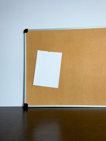 A square, white piece of paper is tacked to a brown corkboard. The board rests on a dark table against a pale wall, waiting for inspiration to strike.の写真素材