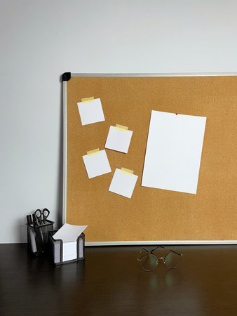 A cork board is mounted on a wall with several blank white cards stuck to it. Also seen are glasses, a pen holder, and notes on a dark wooden desk.の写真素材