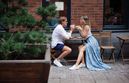 Young trendy Couple Talking at Coffee Shop on a date. Loving couple having Cup of coffee at street restaurant. stylish man and woman full length portrait. cafe table Outdoors coffeehouseの写真素材