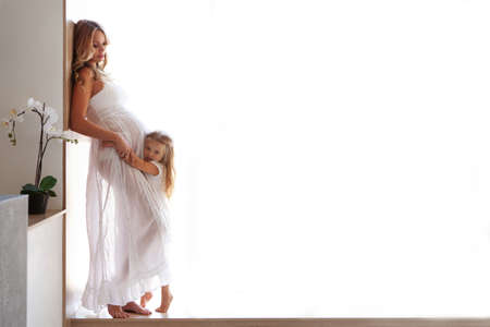 Isolated Pregnant Woman Model with little daughter indoors. Full length portrait with empty space on white background. Little girl listening baby in belly of beautiful mother. Motherhood, pregnancyの写真素材