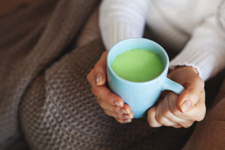 Close up of hands holding blue cup of matcha tea. Model in white sweater and cozy plaid is sitting at home. Depth of field, empty space on blurry beige backgroundの写真素材