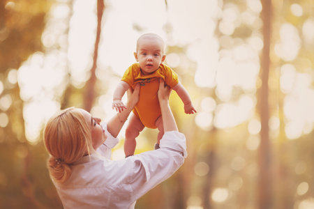 Portrait of a serious baby, which Happy young Mother lifting up high in air, spending and enjoying time together with son. Mom Playing with Little Child in Autumn forest landscape. warm toned photoの写真素材