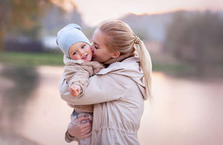 Mom Hugs and Kisses smiling Baby at sunset on nature lake. Blurred background, copy space, beige...