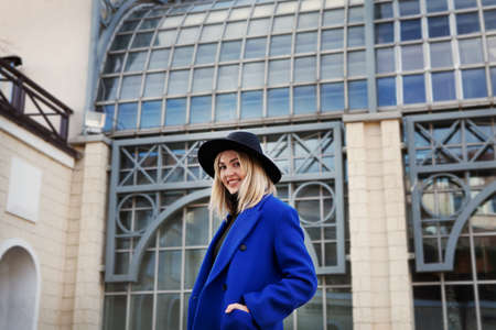 Beautiful young woman in black felt hat and bright blue coat looking at camera and smiling, on background beautiful building. Lifestyle portrait of happy female modelの写真素材
