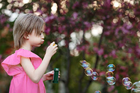 Child girl in pink dress blows soap bubbles in nature against background of flowering treeの写真素材
