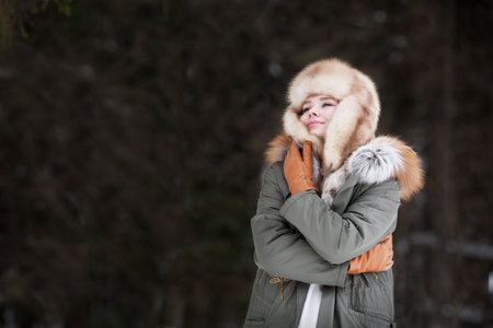 Woman wrap and hug herself on dark background, wearing winter jacket, fur ear flaps hat and leather glove. Cozy portraitの写真素材