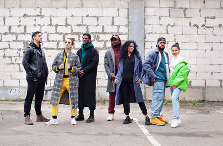 Multiethnic group of young hipsters stands together on street against gray brick wall. Diverse Stylish people in outerwear outdoor, youth cultureの写真素材