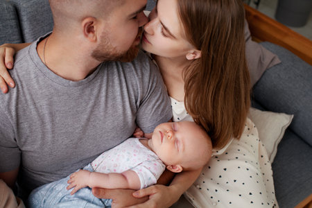 Parents mom and dad kiss and hold newborn sleeping baby in their arms while sitting at home. Family love, close-up portrait. Relationship of man and woman having small childの写真素材