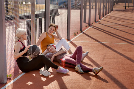 Happy mature women relax on court after playing basketball. Female friends talk, laugh and play sports outdoorの写真素材