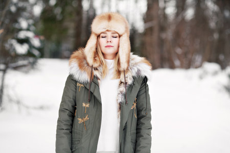 Woman with closed eyes in winter forest. Young female light portrait during relaxation and meditation, wear fur hat and parka jacket, yoga practice outdoors. dreaming girl in natureの写真素材