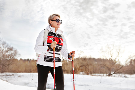 Active mature tourist woman hiking with trekking sticks or cross-country skiing in winter nature against skyの写真素材