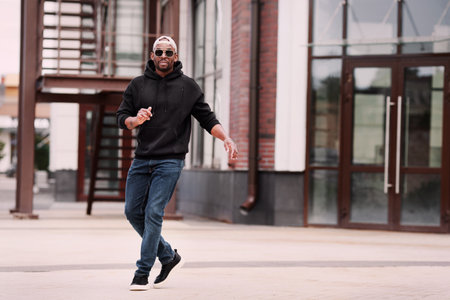 Cool Black man dancing on street of city alone, wears black hoodie, jeans and sneakers, baseball cap and sunglassesの写真素材