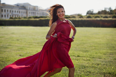 Brunette woman in long, flowing red dress running through park in nature, looking in camera, and smiling. Happy female model with long hair outdoorsの写真素材