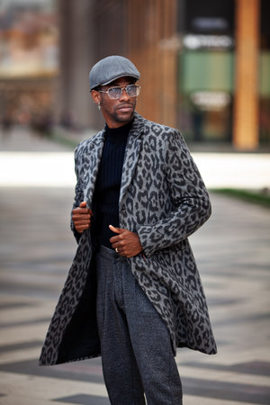 Portrait African American young male in trendy outfit coat with animal print, turtleneck, cap, pants, and glasses, standing on city street. Handsome man, fashion styleの写真素材