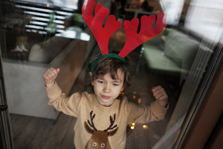 Child in knitted sweater and deer headband stands near window, looks outside. Realistic lifestyle photoの写真素材