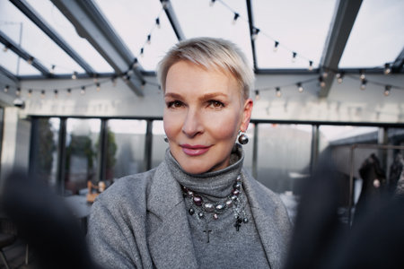 Selfie of beautiful mature blonde woman with short haircut in modern trendy clothes, gray turtleneck, jacket. Close up portrait stylish middle aged female model with brown eyes looking camera indoorの写真素材