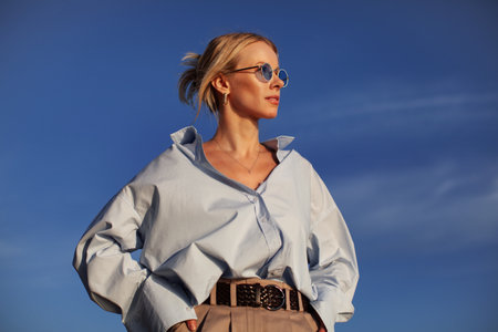 A person in an oversized shirt and sunglasses stands outdoors against the sky. Chic, contemporary,...