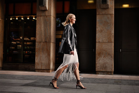 Happy smiling fashionable blonde woman walking city street in chic attire with building in background. Young female model wears white fringed dress, black leather blazer and sandalsの写真素材