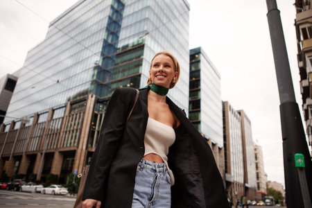 Stylish smiling woman in a modern fashion ensemble, black blazer, top, and jeans, walking confidently along the city streetsの写真素材