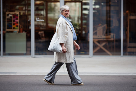 Elderly woman with short gray hair walks down city street wearing fashionable attire pants, coat, sneakers, and tote bag, exuding style and confidence.の写真素材