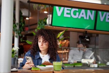 Woman with curly hair seats at vegetarian cafe or restaurant with green smoothie and plant meal on...