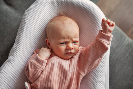 Cute Newborn Baby Girl in Pink Clothing Lying in White Cocoon, Joyfully Making Victory Gesture with Her Hand in Cozy Indoorsの写真素材