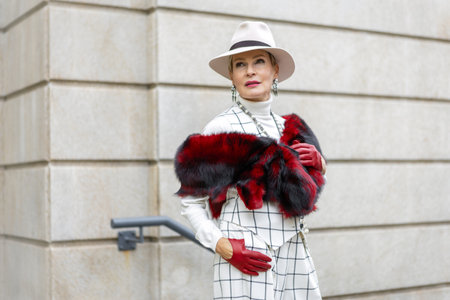 Fashionable Mature Woman Walking Outside in City Dressed in Stylish Checkered White Suit with Fedora Hat, Faux fur Accessories Showcasing Trendy Clothing and Street Styleの写真素材