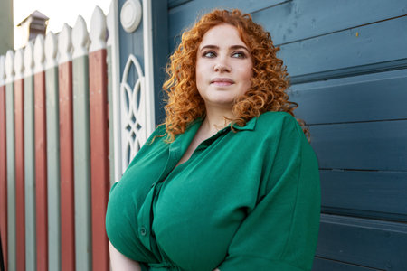 Portrait Plus Size Woman with Curly Red Hair in Green Clothing Against Colorful Blue Wooden Wall Background, Beauty and Confidence Outdoorsの写真素材