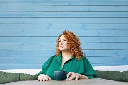 Beautiful Plus-Size Woman with Red Hair Enjoying Cup of Coffee. Curvy Female Model Sitting at Cafe Table in Green Dress Against Bright Colorful Blue Wallの写真素材