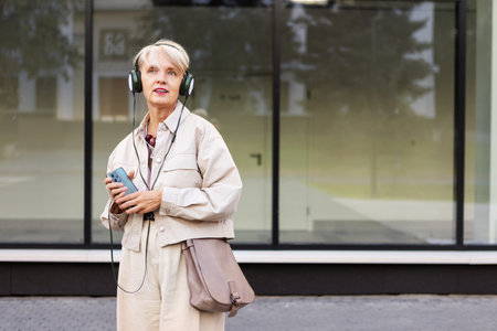 Beautiful Mature Woman with Short Hair Wearing Headphones and Beige Clothing, Walking Outdoors in City While Listening to Music, Podcasts or Audiobookの写真素材
