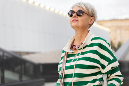 Beautiful Happy Senior Woman with Short hairstyle Wearing Striped Sweater and Sunglasses, Looking Up Against City Background, Confidence and Joyの写真素材