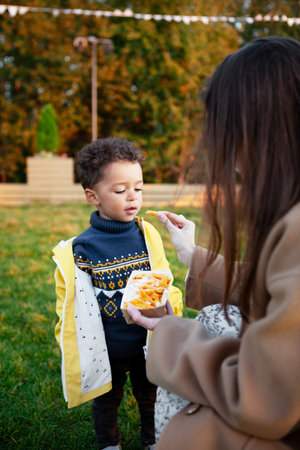 Mother feeding young son French fries at outdoor picnic in park, while child boy in yellow jacket and sweater stands on grass in summer or autumn. Family enjoying snacks and quality time togetherの写真素材