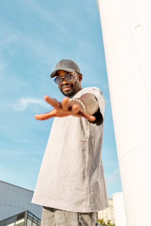 Stylish young African American man wearing casual streetwear, sunglasses and Hat while standing outdoors and reaching out hand against bright blue sky, exuding confidence and positive on sunny dayの写真素材