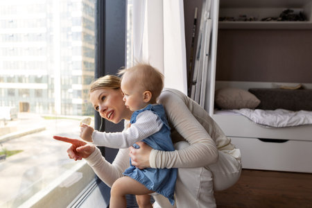 Beautiful mother and little daughter standing by window at home, looking outside, cozy family scene...