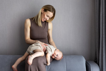 Loving young mother bonding with baby boy at home, sitting on sofa together, enjoying moment in cozy...