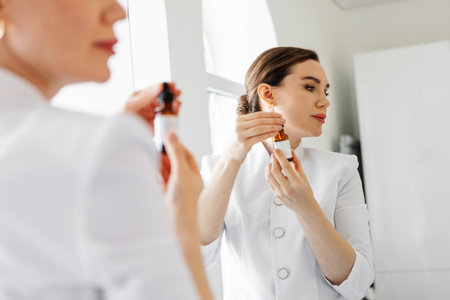 Woman in white coat holding anti-aging serum and dropper near face, in front of mirror in beauty clinic, demonstrating professional skincare product for facial treatment and healthy glowing skinの写真素材