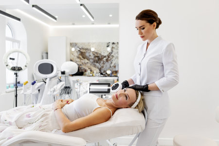 Cosmetologist using skincare equipment for photorejuvenation facial treatment. Mature woman lying on chair in beauty clinic before non-invasive anti-aging procedure with aesthetic laser technologyの写真素材