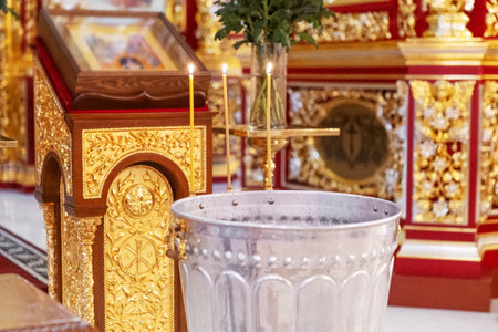 Silver baptismal font inside richly decorated Orthodox church with golden icons and candles. Symbol...