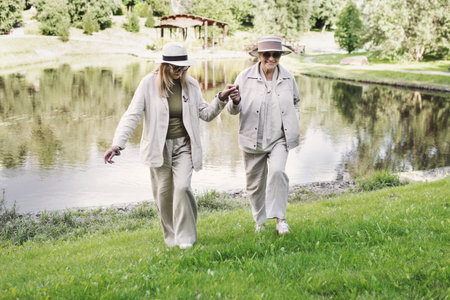 Two senior women friends walking together outdoors in nature near lake, holding hands on green grass, stylish outfits with hats, age positive lifestyle, healthy active leisure, friendship and joyの写真素材