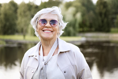 Happy senior woman with white hair smiling outdoors, wearing sunglasses and jacket, enjoying active...
