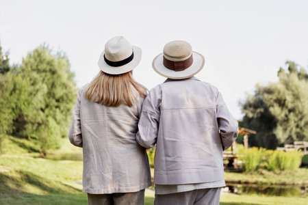 Senior women standing backs outdoors, watching beautiful natural landscape, enjoying peaceful moment, wearing stylish light clothes and straw hats, concept of friendship and active retirement travelの写真素材