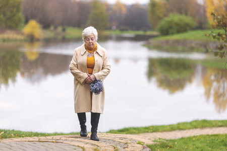 Elderly caucasian woman walking in autumn park alone. Senior lady enjoying peaceful stroll by lake....