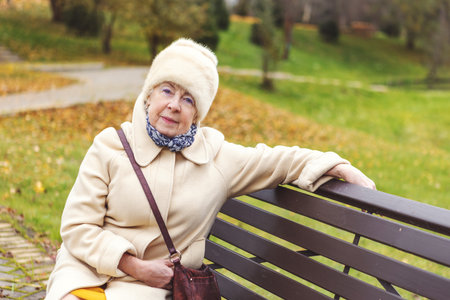 Serious senior woman sitting in autumn park wearing warm coat and scarf. Elderly female looking at camera with calm expression, natural outdoor portrait in peaceful fall natureの写真素材