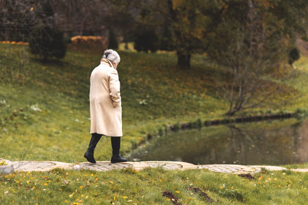 Lonely senior woman walking in autumn park wearing beige coat. Sad elderly female feeling depression, solitude and melancholy outdoors in cold fall nature.の写真素材