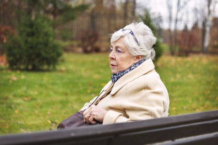 Lonely elderly woman sitting alone on park bench in autumn outdoors. Senior female in warm coat, solitude, sadness, depression and loneliness among older peopleの写真素材