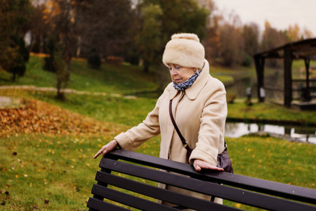 Sad elderly woman standing alone in autumn park, looking down with thoughtful expression. Depressed senior female outdoors surrounded by golden trees and fallen leaves, symbol of loneliness and agingの写真素材