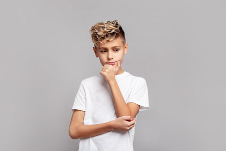 Thoughtful young boy with trendy blond hair touching chin while looking down, wearing white T-shirt and posing in studio, isolated on gray background in modern portraitの写真素材