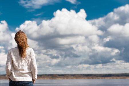 against the background of the river stands a woman with red hair, dressed in a white sweater