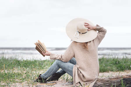 woman reading a book on the beach during a storm, dressed in a warm cardigan and a beige hatの写真素材